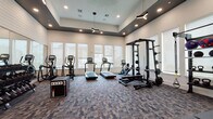 This fitness center is designed to accommodate a full range of workout routines in a spacious, thoughtfully organized environment. The space is entered through double glass doors, which open to reveal a generously proportioned room with notably high ceilings accented by a recessed tray detail finished in a deep gray tone. Modern Y-shaped pendant lights and recessed can lighting provide balanced illumination throughout.

The cardio zone lines the windowed wall, featuring multiple treadmills, elliptical trainers, and a stationary bike, all positioned to take advantage of natural light from a row of large windows fitted with roller shades. The strength training area is equally comprehensive, with a full power rack loaded with weight plates, a multi-function cable machine, an adjustable flat bench, a tiered dumbbell rack spanning a wide weight range, and a freestanding storage unit holding kettlebells and medicine balls.

Large wall mirrors along one side visually expand the room and allow users to monitor their form. The flooring throughout is a durable, multi-tone patterned carpet tile that adds texture while standing up to heavy equipment. The overall ambiance is modern and professional, reflecting a well-equipped athletic club environment.