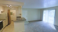 This single-floor residence offers a practical and open layout that connects the kitchen and living areas seamlessly. The kitchen features a peninsula with a dark laminate countertop and an integrated sink, providing a natural divider between the cooking and living spaces. White cabinetry lines the kitchen walls, offering ample storage, and a fire extinguisher is conveniently mounted nearby. The transition from tile flooring in the kitchen to plush wall-to-wall carpeting marks the entry into the spacious living area. The living room is bright and open, with neutral walls and a textured ceiling that add a clean, understated aesthetic. A full-height sliding glass door floods the space with natural light and provides access to an outdoor area, enhancing the sense of openness. A solid interior door is centrally positioned along the back wall, likely leading to additional private rooms. Soft, neutral tones throughout the space create a calm and welcoming atmosphere, making it a versatile canvas ready for personalization.