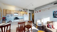 This single-floor space showcases an open-concept layout that seamlessly integrates the kitchen and living areas. The design draws attention with its exposed silver ductwork running along the ceiling, lending an industrial aesthetic that contrasts with the otherwise light and airy interior. White-painted structural beams further define the ceiling line while maintaining an open feel throughout.

The kitchen is equipped with light maple cabinetry, a white refrigerator, microwave, and standard appliances including a coffee maker and stovetop. A built-in sink with overhead cabinet storage maximizes functionality. A peninsula counter with bar-height seating accommodates two wooden chairs, creating a casual dining or workspace option directly connected to the kitchen.

The living area is bathed in natural light and features neutral-toned flooring. A doorway visible from the main space leads to what appears to be a bathroom, adding to the unit's practical layout.

The overall ambiance is bright and functional, with the industrial ceiling elements balanced by warm wooden furniture accents and decorative string lights, creating a comfortable and livable environment.
