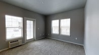 This single-floor space presents a clean, contemporary aesthetic centered around a well-proportioned main room finished in a consistent neutral grey palette. The walls carry a smooth, medium-grey tone that pairs cohesively with the white trim and baseboards running throughout, lending the interior a polished, modern feel.

The room is finished with wall-to-wall carpet in a light, textured grey-beige tone, providing a soft underfoot surface that contributes to the overall warmth of the space. Natural light is a notable feature, introduced through two side-by-side grid-pane windows positioned along one wall, as well as a full-length glass-paneled exterior door with a decorative privacy window inset, both fitted with horizontal blinds for light control and privacy.

The layout of the room is open and versatile, offering ample space for a variety of furniture arrangements. The smooth, flat ceiling adds to the clean, uncluttered character of the interior. Electrical outlets are conveniently distributed along the walls, and a smoke detector is centrally mounted on the ceiling. Overall, the space offers a neutral, move-in-ready environment with a bright and airy atmosphere.