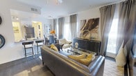 This single-floor residence showcases a thoughtfully designed open-concept layout that connects the living, dining, and kitchen areas into one cohesive space. The living room serves as the focal point, anchored by a substantial gray leather sectional sofa accented with gold and yellow throw pillows, complementing the warm, contemporary aesthetic throughout. A sleek glass coffee table sits at the center, while a dark console cabinet displaying curated decorative art adds visual depth to the space. Large windows dressed in flowing gray curtains allow natural light to filter in, enhancing the bright, airy atmosphere. Rich dark hardwood flooring runs throughout, providing a grounding contrast to the white walls and light ceiling with crown molding details. The dining area, visible in the background, features a round table surrounded by upholstered chairs, with warm pendant lighting overhead contributing to an intimate ambiance. Adjacent to the dining space, the kitchen area is equipped with a breakfast bar and counter seating, ideal for casual dining. A large abstract artwork displayed prominently on one wall adds a bold artistic statement, tying together the modern and sophisticated character of the entire space.