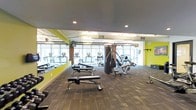 This contemporary fitness center is designed to support a well-rounded workout experience within a single, open-plan floor. The space is finished with durable patterned carpet flooring and features a bold color scheme, with vibrant lime-green accent walls contrasting against a sleek dark ceiling — giving the gym a modern, energetic atmosphere.

Natural light pours in through a wide bank of floor-to-ceiling windows along one wall, providing an open and airy feel while offering views to the outdoors. The windows are complemented by recessed and overhead fluorescent lighting throughout, ensuring the space remains well-lit at all hours.

The fitness floor is thoughtfully laid out with dedicated zones for different types of training. A row of cardio machines — including treadmills and ellipticals — is positioned along the windowed wall, allowing users to enjoy the natural light during their workouts. The central area houses a multi-station cable and strength machine, ideal for a variety of resistance exercises. Adjustable weight benches and free weights are arranged nearby, offering a complete strength training setup.

Wall-mounted accessories, including pull-up and stretching bars, are also visible, rounding out the functional offerings of this well-appointed fitness facility.