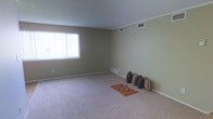 This single-floor space offers a welcoming and functional layout designed with simplicity and comfort in mind. The main living area is a standout feature, boasting plush wall-to-wall carpeting in a neutral tone that adds warmth underfoot and complements the soft sage-green walls. A large window along one wall floods the room with abundant natural light, creating a bright and airy atmosphere throughout the day. Crown molding trims the ceiling, lending a subtle touch of architectural detail, while multiple ceiling vents ensure efficient climate control throughout the space. The room's generous proportions provide ample flexibility for a variety of furniture arrangements and lifestyle needs. Clean white baseboards frame the perimeter of the room, adding a crisp contrast to the muted wall color. The overall ambiance is calm and neutral, offering a versatile blank canvas that is well-suited for customization. Whether used as a primary living space or a relaxed gathering area, this room combines practicality with an understated aesthetic that is easy to build upon.