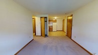Located at 144 Dunbar in Manchester, New Hampshire, this single-floor unit offers a straightforward and functional layout designed for comfortable everyday living. The main living area is carpeted in a neutral tone, providing a clean and understated foundation that makes the space feel open and adaptable to various furnishing styles. Smooth walls with warm wood-toned baseboards and door trim run throughout, lending a cohesive and traditional aesthetic. The kitchen is positioned toward the rear of the main living area and features wooden cabinetry, a white refrigerator, and a countertop workspace, keeping essential amenities within easy reach. Multiple interior doors lead to additional rooms and spaces, suggesting a well-organized floor plan with defined areas for sleeping, storage, and utility. A closet with a bi-fold door is visible near the main living space, offering convenient built-in storage. The front entry door provides direct access to the unit and is framed in matching warm wood tones consistent with the rest of the interior. Ceiling-mounted lighting provides consistent illumination throughout. Overall, the space presents a clean, practical interior that prioritizes function and ease of use.
