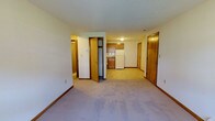 Located at 144 Dunbar in Manchester, New Hampshire, this single-floor unit offers a straightforward and functional layout designed for comfortable everyday living. The main living area is carpeted in a neutral tone, providing a clean and understated foundation that makes the space feel open and adaptable to various furnishing styles. Smooth walls with warm wood-toned baseboards and door trim run throughout, lending a cohesive and traditional aesthetic. The kitchen is positioned toward the rear of the main living area and features wooden cabinetry, a white refrigerator, and a countertop workspace, keeping essential amenities within easy reach. Multiple interior doors lead to additional rooms and spaces, suggesting a well-organized floor plan with defined areas for sleeping, storage, and utility. A closet with a bi-fold door is visible near the main living space, offering convenient built-in storage. The front entry door provides direct access to the unit and is framed in matching warm wood tones consistent with the rest of the interior. Ceiling-mounted lighting provides consistent illumination throughout. Overall, the space presents a clean, practical interior that prioritizes function and ease of use.