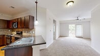 Located at 10505 W 134th St in Overland Park, KS, this one-bedroom, one-bathroom residence offers a well-designed layout that maximizes comfort and functionality. The open-concept kitchen is a standout feature, boasting rich dark-stained cabinetry, dark granite countertops, a tile backsplash, and a full suite of stainless steel appliances including a refrigerator, range, microwave, and dishwasher. A peninsula with a double-basin sink and pendant lighting creates a natural divide between the kitchen and the adjoining living area, which is enhanced by crown molding and a ceiling fan. Plank hardwood-style flooring runs through the kitchen, while plush carpet extends throughout the living room and bedroom. The living area is bright and open, with a sliding glass door providing access to an outdoor space and filling the room with natural light. The generously sized bedroom features dual windows, crown molding, a ceiling fan, and a closet with double doors. The bathroom is well-appointed with a dark granite vanity, rich wood cabinetry, a tiled tub-shower combination, and hardwood-style flooring. Crown molding is carried consistently throughout the home, adding a refined, cohesive touch to the overall aesthetic.