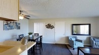 This single-floor residence offers an open-concept layout that seamlessly integrates the dining and living areas into one cohesive space. The interior is finished with dark hardwood-style flooring throughout, which contrasts neatly against the light, neutral-toned walls. A textured popcorn ceiling adds a classic touch overhead, while a ceiling fan with a light fixture provides both airflow and ambient lighting to the dining area.

The dining area accommodates a dark wood table and matching chairs, positioned near a window fitted with vertical blinds that allow for adjustable natural light. Decorative wall art, including a sculptural fish school piece, adds character to the space. Adjacent to the dining area, the living space features a patterned accent chair and an area rug, creating a defined seating zone. A framed mirror mounted on the wall enhances the sense of openness. The front entry door is centrally located along the main wall, offering straightforward access. Overall, the space presents a functional and modestly styled interior suited for everyday living.