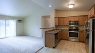 This single-story home showcases an open-concept layout that seamlessly connects the kitchen and living area, creating a cohesive and functional living space. The kitchen is well-appointed with warm honey-toned wood cabinetry that provides ample storage, complemented by dark granite countertops that add a refined touch. Stainless steel appliances, including a gas range, built-in microwave, and dishwasher, lend a modern, polished aesthetic to the cooking space. A peninsula with a built-in sink and matching granite surface extends into the open area, offering additional workspace and serving as a natural divider between the kitchen and the adjacent living space. The kitchen features tile flooring, while the living area transitions to plush wall-to-wall carpeting, distinguishing the two zones. Vaulted ceilings in the living area contribute to an airy, spacious feel, while recessed lighting in the kitchen and a flush-mount ceiling fixture ensure the space is well-lit. The neutral wall tones throughout create a clean, versatile backdrop adaptable to a variety of interior styles.