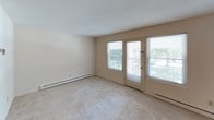 This single-floor, one-bedroom, one-bathroom unit is thoughtfully laid out to maximize functionality and comfort. The living area features neutral-toned carpet, a ceiling fan with light fixture, and an open passthrough connecting to the kitchen, creating a cohesive and airy feel. A private exterior door flanked by large windows floods the living space with natural light and provides convenient outdoor access. The kitchen is equipped with dark wood cabinetry offering ample storage, light countertops, a coil-element range, dishwasher, and a drop ceiling with panel lighting, giving it a practical and organized workspace. The bedroom is a comfortable, carpeted retreat with a window overlooking greenery and baseboard heating for year-round comfort. A spacious walk-in closet off the bedroom features a full-width shelf and hanging rod, providing generous storage. A utility area conveniently houses a stacked washer and dryer unit alongside built-in shelving. The full bathroom includes a tiled tub and shower surround with grab bars, a vanity with mirror, and a drop ceiling, maintaining a clean, functional aesthetic throughout. Baseboard heating is present in multiple rooms, ensuring consistent warmth across the unit.