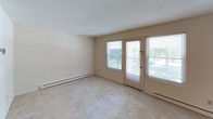 This single-floor, one-bedroom, one-bathroom unit is thoughtfully laid out to maximize functionality and comfort. The living area features neutral-toned carpet, a ceiling fan with light fixture, and an open passthrough connecting to the kitchen, creating a cohesive and airy feel. A private exterior door flanked by large windows floods the living space with natural light and provides convenient outdoor access. The kitchen is equipped with dark wood cabinetry offering ample storage, light countertops, a coil-element range, dishwasher, and a drop ceiling with panel lighting, giving it a practical and organized workspace. The bedroom is a comfortable, carpeted retreat with a window overlooking greenery and baseboard heating for year-round comfort. A spacious walk-in closet off the bedroom features a full-width shelf and hanging rod, providing generous storage. A utility area conveniently houses a stacked washer and dryer unit alongside built-in shelving. The full bathroom includes a tiled tub and shower surround with grab bars, a vanity with mirror, and a drop ceiling, maintaining a clean, functional aesthetic throughout. Baseboard heating is present in multiple rooms, ensuring consistent warmth across the unit.