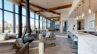 Located at 320 W 13th Ave in Denver, Colorado, this single-level amenity lounge is a thoughtfully designed communal space that blends industrial character with contemporary finishes. Floor-to-ceiling windows line the perimeter, flooding the interior with natural light and framing sweeping views of the Denver skyline, making the cityscape a defining feature of the space.

Exposed wood beams cross the ceiling, complementing the raw texture of an exposed brick wall that anchors one side of the room. A striking suspended fireplace serves as a focal point within the lounge seating area, which is furnished with plush sofas and accent chairs arranged around a round wood coffee table, creating an inviting atmosphere for conversation and relaxation.

The game area features a pool table positioned beneath pendant lighting, adding an entertainment-focused dimension to the space. Adjacent to it, a well-appointed wet bar commands attention with its dramatic marble countertop and waterfall edge, teal mosaic tile accent wall, dark navy cabinetry with brass hardware, stainless steel appliances, and a wall-mounted television. Pendant lighting clusters above the bar, enhancing the upscale ambiance. Throughout, the design harmonizes natural materials, bold color accents, and panoramic urban views into a cohesive, vibrant gathering space.