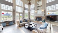 Located at 1 Applewood Drive in Perkasie, Pennsylvania, this single-floor community space offers a generous and thoughtfully designed interior. The centerpiece is a dramatic living room boasting soaring two-story ceilings lined with large transom windows that flood the space with natural light. A striking floor-to-ceiling stone fireplace anchors the room, complemented by an elegant chandelier, hardwood flooring, and neutral tones that create a warm, sophisticated ambiance. The open layout transitions seamlessly through an arched hallway, connecting the main gathering area to the additional rooms. The dining room features classic wainscoting with beadboard paneling, crown molding, patterned carpet, and a decorative ceiling fan, providing a comfortable and functional space for group meals or meetings. A wall-mounted display screen adds a modern, versatile touch. Outside, a spacious wraparound deck with white columned railings extends the living space outdoors, offering views of lush greenery and a nearby pool. The exterior showcases attractive stone veneer accents and French doors that open onto the deck, enhancing the connection between indoor and outdoor spaces.