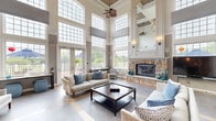 Located at 1 Applewood Drive in Perkasie, Pennsylvania, this single-floor community space offers a generous and thoughtfully designed interior. The centerpiece is a dramatic living room boasting soaring two-story ceilings lined with large transom windows that flood the space with natural light. A striking floor-to-ceiling stone fireplace anchors the room, complemented by an elegant chandelier, hardwood flooring, and neutral tones that create a warm, sophisticated ambiance. The open layout transitions seamlessly through an arched hallway, connecting the main gathering area to the additional rooms. The dining room features classic wainscoting with beadboard paneling, crown molding, patterned carpet, and a decorative ceiling fan, providing a comfortable and functional space for group meals or meetings. A wall-mounted display screen adds a modern, versatile touch. Outside, a spacious wraparound deck with white columned railings extends the living space outdoors, offering views of lush greenery and a nearby pool. The exterior showcases attractive stone veneer accents and French doors that open onto the deck, enhancing the connection between indoor and outdoor spaces.