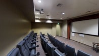 This single-floor screening room is purpose-built for an immersive viewing experience, combining functional design with a refined cinematic atmosphere. Rows of padded, theater-style seats are arranged to ensure clear sightlines to the front of the room, with each seat equipped with individual armrests and built-in cup holders for added comfort during extended viewings. A ceiling-mounted projector is centrally positioned overhead, directing content onto a large projection screen at the front of the room, framed by warm, rust-toned drape curtains that add a classic theatrical touch. The walls are finished in a muted, neutral tone, contributing to the room's focused and distraction-free environment. Recessed lighting is thoughtfully integrated into the ceiling, offering controlled ambient illumination that complements the room's presentation function. Ventilation is provided through ceiling-mounted HVAC vents, ensuring a comfortable climate throughout. The flooring features a durable tile finish suited to the room's high-use nature. This space is well-suited for private film screenings, corporate presentations, educational sessions, or any event requiring a dedicated and well-equipped viewing environment.
