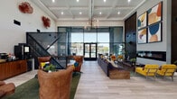 This single-story community clubhouse is designed with a contemporary aesthetic, combining open-plan living with functional, defined spaces. The main lounge area commands attention with its soaring coffered ceilings, light wood-look flooring, and dramatic floor-to-ceiling glass walls that frame outdoor views of a pool area and lush landscaping, flooding the interior with natural light. A striking globe chandelier anchors the lounge, which is furnished with a large sectional sofa, cognac leather armchairs, and bold yellow velvet seating arranged around a sculptural wood coffee table. A linear fireplace set into a dark wood-paneled accent wall serves as a focal point, topped with vibrant abstract artwork. Decorative coral-toned wall sculptures add artistic character throughout.

A glass-enclosed conference room with a geometric-patterned feature wall and ergonomic seating provides a dedicated workspace. The leasing office area, also enclosed in full-height glass, houses multiple wooden desks, blue upholstered chairs, and private meeting rooms bathed in natural light from tall windows overlooking palm-lined surroundings. The cohesive design blends warm wood tones, bold colors, and modern architectural elements into a welcoming, multifunctional environment.