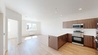 This contemporary 2-bedroom, 1-bathroom unit is thoughtfully designed with a clean, modern aesthetic throughout. The open-concept kitchen and living area serves as the heart of the home, featuring light hardwood-style flooring, recessed lighting, and a large window that floods the living space with natural light. The kitchen is well-appointed with rich wood-grain cabinetry extending to the ceiling, a full suite of stainless steel appliances including a refrigerator, electric range, built-in microwave, and dishwasher, complemented by crisp white countertops and a peninsula that provides additional prep and seating space. Both bedrooms are bright and comfortable, each featuring large windows with views of the surrounding neighborhood, light hardwood flooring, and wall-mounted heating units. The primary bedroom includes a ceiling fan for added comfort. The full bathroom is finished with a wood-toned vanity, white countertop, large mirror, and hardwood-style flooring, with grab bars installed for accessibility. Clean white walls and a consistent flooring palette unify the space, giving the unit a cohesive, move-in-ready feel.