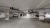 This underground parking facility encompasses a generous single-level footprint, providing substantial capacity for vehicle storage. The structure is defined by a concrete ceiling from which an exposed network of pipes, conduits, and mechanical systems runs throughout, supported by regularly spaced load-bearing columns painted white with distinctive yellow accent bands for visibility and safety.

The floor consists of poured concrete, with individual parking stalls delineated by yellow painted lines. Designated accessible parking spaces are clearly identified with standard blue signage posted on the walls and columns throughout the facility. The interior is illuminated by rows of fluorescent strip lighting mounted to the ceiling, ensuring adequate visibility across the entire space.

The white-painted perimeter walls contribute to the brightness of the environment. Multiple access points are present, including doors that lead to stairwells and elevator lobbies, facilitating convenient entry and exit for users. The layout, as visible in the 3D overview, features an irregular footprint with multiple interconnected sections, maximizing the available parking area. The facility is fully enclosed, offering protected, covered parking suitable for a variety of vehicles.