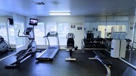 This fitness center is a compact yet fully functional workout space designed to accommodate a variety of exercise routines. The room features durable rubber flooring throughout, providing a safe and supportive surface for both cardio and strength training activities. Natural light filters in through multiple windows fitted with white horizontal blinds, creating a bright and energizing atmosphere complemented by overhead fluorescent lighting. A wall-mounted flat-screen TV adds an entertainment option for those engaging in longer cardio sessions. The cardio section includes a treadmill and an elliptical machine, while a stationary bike serves as an additional low-impact option. A full wall-length mirror runs along one side of the room, enhancing the sense of space and allowing users to monitor their form during workouts. The strength training area features a well-stocked dumbbell rack with a wide range of weights neatly organized on a multi-tiered stand. Informational and safety signage is posted on the walls, contributing to a structured and professional environment. The space is thoughtfully arranged to maximize usability within its footprint, making it a practical and convenient fitness facility.