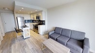 This well-designed single-floor space presents a seamless open-concept layout that connects the kitchen and living area, creating a cohesive and functional environment. The kitchen is outfitted with stainless steel appliances, including a refrigerator, range, and microwave, complemented by crisp white cabinetry and a stylish dark tile backsplash that adds a modern contrast. A stainless steel sink sits beneath recessed lighting, and the counter space offers ample room for meal preparation. A portable kitchen cart provides additional storage and flexibility. The living area flows naturally from the kitchen, anchored by a contemporary gray sofa and a minimalist wood-and-metal coffee table, creating a relaxed and inviting atmosphere. Light wood-toned flooring runs throughout the space, lending warmth and visual continuity to the overall design. The clean white walls and recessed ceiling lights contribute to a bright and airy ambiance. The overall aesthetic is modern and understated, with a neutral palette that offers a versatile backdrop. The thoughtful layout maximizes the available space, making it both practical and comfortable for everyday living.