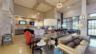 This clubhouse delivers a sophisticated and welcoming atmosphere through a thoughtfully curated blend of modern and industrial design elements. The grand entrance foyer sets the tone immediately, featuring soaring double-height ceilings, polished concrete floors, floor-to-ceiling black-framed glass doors and transom windows, and stacked stone accent columns that flood the space with natural light while maintaining an upscale aesthetic.

The main lounge area is expansive and elegantly furnished, offering a mix of plush leather sofas, vibrant coral accent chairs, and sleek side tables. Statement pendant lighting and a coffered ceiling add architectural depth, while large windows maintain a bright, airy ambiance. A striking built-in linear fireplace clad in mosaic tile serves as a central focal point, flanked by a generous built-in bench seat dressed with bold red and patterned throw pillows. Above the fireplace, a wall-mounted flat-screen TV and an artful arrangement of decorative metallic discs on the stacked stone wall add visual interest and warmth.

Adjacent to the lounge, a leasing and amenity bar area features a clean white countertop, modern seating, and a reclaimed wood accent wall hosting multiple mounted screens and decorative golden sunburst sculptures. A beverage station with a coffee machine and mini-refrigerator rounds out this functional yet stylish community hub.