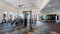 Located at 1124 Shoreview Cir in Casselberry, Florida, this single-floor fitness center is a well-appointed facility designed to support a comprehensive workout experience. The space is defined by its elevated tray ceilings with warm accent lighting and modern ceiling fans, creating an open and energizing atmosphere throughout.

The main workout floor is covered in a stylish multi-toned carpet tile in shades of blue, teal, and taupe, providing both comfort and visual interest. Large windows along the perimeter flood the space with natural light and offer views of the surrounding landscaping, contributing to an uplifting environment.

The cardio zone features a row of treadmills, ellipticals, and stationary cycling bikes equipped with individual digital screens, alongside wall-mounted televisions. The free weights area is lined with floor-to-ceiling mirrors that visually expand the space, and includes a full dumbbell rack, adjustable benches, and barbell stations. A cable and functional training machine occupies a central position on the main floor.

A dedicated functional training room offers open floor space, battle ropes, medicine balls, a punching bag, and additional resistance equipment — ideal for dynamic or group workouts. The entryway features clean tile flooring, glass-paneled walls, and double doors that open to the exterior. Hydration amenities include a dual water fountain station and a small refreshment counter.