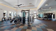 Located at 1124 Shoreview Cir in Casselberry, Florida, this single-floor fitness center is a well-appointed facility designed to support a comprehensive workout experience. The space is defined by its elevated tray ceilings with warm accent lighting and modern ceiling fans, creating an open and energizing atmosphere throughout.

The main workout floor is covered in a stylish multi-toned carpet tile in shades of blue, teal, and taupe, providing both comfort and visual interest. Large windows along the perimeter flood the space with natural light and offer views of the surrounding landscaping, contributing to an uplifting environment.

The cardio zone features a row of treadmills, ellipticals, and stationary cycling bikes equipped with individual digital screens, alongside wall-mounted televisions. The free weights area is lined with floor-to-ceiling mirrors that visually expand the space, and includes a full dumbbell rack, adjustable benches, and barbell stations. A cable and functional training machine occupies a central position on the main floor.

A dedicated functional training room offers open floor space, battle ropes, medicine balls, a punching bag, and additional resistance equipment — ideal for dynamic or group workouts. The entryway features clean tile flooring, glass-paneled walls, and double doors that open to the exterior. Hydration amenities include a dual water fountain station and a small refreshment counter.