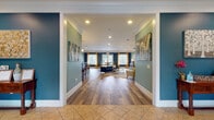 This single-story clubhouse in Brunswick, Georgia offers a thoughtfully designed community hub with a variety of amenity spaces finished in a cohesive coastal-inspired color palette of teal, sage green, and navy blue, accented by crisp white crown molding and trim throughout. The welcoming entry foyer features tile flooring, a built-in bench with decorative pillows, potted plants, and a reception window, setting an inviting tone from the moment of arrival. The main lounge area transitions into rich wood-look flooring and showcases a white fireplace mantel with a wall-mounted TV, yellow accent chairs, and a decorative area rug — an ideal space for relaxed socializing. Adjacent is a dedicated billiards room with a full-size pool table and pendant lighting overhead. A bar-height counter with a granite top and red metal stools provides a casual gathering spot, complemented by a nearby coffee and refreshment station. The dining area features a solid wood table and chairs, with French doors opening directly to an outdoor pool area, flooding the space with natural light. A separate lounge nook with deep red walls, a tufted sofa, and leather armchairs offers a more intimate retreat. The fitness center is equipped with cardio and strength training machines and features rubber flooring, windows for natural light, and a dedicated exterior exit.