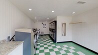 This single-floor shared laundry facility is designed for efficiency and ease of use, offering a clean, well-organized environment for residents. The room is outfitted with a row of front-loading washing machines, including both standard and commercial-grade stainless steel units, positioned along one wall. On the opposite side, a bank of stacked dryers provides ample drying capacity to accommodate multiple users simultaneously. A built-in payment kiosk is conveniently located near the dryers for easy access.

The space features a generous folding and sorting area, with a granite-topped counter running along one wall, offering ample workspace. A recessed ironing station with a built-in iron and board, along with a hanging rod, provides added convenience for garment care. The flooring throughout is a classic green and white checkered tile that adds a retro, clean aesthetic to the space.

Recessed lighting illuminates the interior evenly, contributing to a bright and functional atmosphere. A large glass door and wide window at the entrance flood the room with natural light and provide a clear view of the surrounding outdoor parking area. White beadboard-style wall paneling and neutral walls maintain a tidy, welcoming appearance throughout the facility.