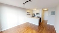 This well-appointed single-floor unit offers a thoughtfully designed open-concept layout that seamlessly connects the kitchen and living areas. Rich hardwood flooring runs throughout the main living spaces, adding warmth and continuity to the modern aesthetic.

The kitchen is a standout feature, equipped with white shaker-style upper cabinets, dark lower cabinetry, and a classic white subway tile backsplash. Stainless steel appliances — including a refrigerator, electric range, and over-the-range microwave — complement the quartz countertops and peninsula island, which provides additional prep space and casual seating. Open wooden shelves mounted on the wall add both style and functionality.

The living area is bathed in natural light, enhanced by a floor-to-ceiling glass door that opens to a private balcony with views of the surrounding neighborhood. Track lighting is installed throughout, offering adjustable, contemporary illumination. A ceiling-mounted HVAC unit ensures efficient climate control year-round.

The bathroom is finished with large-format neutral subway tile surrounding the full soaking tub and shower combination. A floating wood vanity topped with a speckled quartz countertop and an undermount sink pairs with a modern chrome faucet, while a backlit LED mirror adds a polished, spa-like touch. The overall ambiance is clean, modern, and move-in ready.