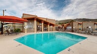 Located at 1901 Brown St in El Paso, Texas, this outdoor pool area offers a well-appointed recreational space set against the striking backdrop of the Franklin Mountains. The generously sized swimming pool features clearly marked shallow and deep ends, with convenient stainless steel entry rails providing easy access. The surrounding concrete deck is outfitted with multiple lounge chairs, offering ample space for relaxation in the abundant West Texas sunshine.

A covered colonnade with distinctive red-trimmed wood beams runs along one side of the pool area, providing shaded seating and shelter. Bold red shade umbrellas and pergola structures are positioned around the deck, offering additional sun protection throughout the day. Decorative stone landscaping elements, potted plants, and globe accent lighting add character and warmth to the environment.

The pool area is fully enclosed by a wrought iron security fence, with a safety life preserver ring mounted and visible on the perimeter. The surrounding terrain — including the rugged mountain hillside — is visible beyond the fence, lending a dramatic and open feel to the space. The combination of Southwestern architectural details, open skies, and panoramic mountain views creates an inviting and visually distinctive outdoor retreat.