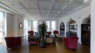 This single-floor space makes an immediate impression with its elegantly detailed coffered ceiling, which adds a sense of architectural refinement and grandeur to the interior. Large windows line the perimeter, flooding the room with natural light and offering open views of the surrounding landscape, enhancing the bright and airy atmosphere throughout.

The lounge area is furnished with an eclectic and vibrant mix of seating, including a patchwork sofa in a rich variety of patterns and colors, complemented by deep crimson velvet armchairs that add warmth and a touch of luxury. A tall, lush indoor plant serves as a natural focal point, bringing life and greenness to the space.

A decorative Moorish-arched alcove frames a built-in media unit and shelving, adding a distinctive architectural accent that blends Eastern and classical influences. The warm hardwood parquet flooring ties the space together, providing an elegant contrast to the white walls and ceiling.

The overall ambiance is one of sophisticated eclecticism — a thoughtfully curated environment that balances bold design choices with a welcoming, comfortable atmosphere suited for relaxation or social gatherings.