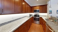 This single-story clubhouse offers a thoughtfully designed gathering space suited for both casual socializing and organized events. The main lounge area serves as a warm focal point, centered around a classic fireplace with a crisp white mantel, flanked by built-in shelving and wall sconces. A wall-mounted television sits above the fireplace, and large windows with French doors fill the room with natural light while offering views of surrounding greenery. Hardwood flooring, a patterned area rug, and comfortable seating complete the inviting atmosphere.

The clubhouse includes two kitchen areas. One features rich cherry wood cabinetry, granite countertops, stainless steel appliances, and generous counter space. The second kitchen is outfitted with a large peninsula with pendant lighting overhead, full-size refrigerator, and matching cabinetry — well suited for catering or community events.

A spacious open room with soaring vaulted ceilings and a skylight provides an airy, flexible area for gatherings or activities. French doors lead to an outdoor area, while framed artwork and warm hardwood floors lend the space a polished yet welcoming character throughout.