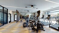 This single-floor fitness center is thoughtfully designed to accommodate a wide range of workout needs within a cohesive, modern aesthetic. The main cardio area is expansive, lined with Matrix treadmills, ellipticals, and a rowing machine, all equipped with individual screens. Full-length mirrors along one wall visually extend the space, while large windows and floor-to-ceiling glass panels invite abundant natural light and outdoor views of surrounding greenery. The distinctive geometric hexagonal flooring in neutral tones of tan, gray, and ivory adds a refined visual element throughout the main floor.

A striking slatted wood partition wall serves as an architectural focal point, dividing the main workout area from additional training zones. Beyond it, a dedicated strength area offers a cable machine system, free weights, benches, and colorful functional training equipment including medicine balls and resistance tools displayed on organized racks.

A separate enclosed studio, accessible through black-framed glass doors, houses spinning bikes alongside bold abstract blue artwork, creating an energizing yet polished atmosphere. Linear LED lighting runs along the ceilings throughout, complementing the clean, contemporary design and ensuring consistently well-lit workout conditions across all zones.