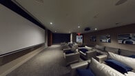 This private screening room is designed with a refined, cinematic aesthetic that prioritizes comfort and visual performance. A large wall-mounted projection screen anchors the front of the room, paired with a ceiling-mounted projector to deliver an immersive viewing experience. Recessed lighting is thoughtfully distributed across the smooth white ceiling, providing subtle ambient illumination that complements the room's dark, moody palette.

The seating arrangement consists of multiple generously sized grey leather sofas and lounge chairs, accented with navy blue throw pillows, arranged to ensure clear sightlines to the screen from every position. Small dark side tables are positioned between seating clusters for added convenience.

The walls feature a combination of deep charcoal textured panels and warm taupe finishes, contributing to the room's acoustic and visual sophistication. A striking wood-paneled accent element adds warmth near the screen wall. Along the side wall, a curated series of bold blue-and-white abstract artworks adds a gallery-like quality to the space. Plush, patterned carpeting in neutral tones covers the floor throughout, enhancing sound absorption and overall comfort. The result is a polished, theatre-inspired space ideal for private screenings and entertainment.
