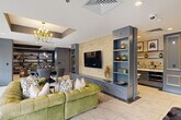 Located in the heart of Midtown Atlanta at 811 Juniper St NE, this elegantly designed living room serves as a refined communal gathering space. The single-floor layout makes excellent use of its footprint, offering distinct functional zones that flow seamlessly into one another.

The space is anchored by large floor-to-ceiling windows and glass doors that flood the interior with natural light while providing direct access to an outdoor terrace with views of the surrounding greenery. Polished marble-style tile flooring runs throughout, lending a clean, upscale foundation to the design.

A tufted green leather sectional sofa and a round tufted ottoman define the primary lounge area, complemented by a coffered tray ceiling adorned with a gold chandelier. A textured, pearl-toned accent wall frames a large mounted flat-screen TV flanked by built-in slate-blue shelving units.

Adjacent dining and bar areas add versatility — a marble counter with brass-accented bar stools and decorative framed panels creates an intimate bar setting, while a separate dining nook features deep navy built-in shelving, pendant lighting, and a dark round table. A cozy library-style corner with floor-to-ceiling shelving and a built-in sofa adds a quiet retreat.

A fireplace with a classic mantel and curated artwork, including a bold dragon canvas, enhances the eclectic yet sophisticated ambiance throughout.