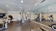 This dedicated fitness center offers a well-organized single-room layout designed to support a complete workout experience. The space features durable, dark rubberized flooring that provides both traction and cushioning underfoot, ideal for high-impact exercise. Recessed lighting is evenly distributed across the white ceiling, ensuring bright, consistent illumination throughout the room. A prominent floor-to-ceiling mirror spans one full wall, visually expanding the space and allowing users to monitor their form during exercise. The strength training area is anchored by a multi-function cable machine and additional resistance equipment, providing a range of upper and lower body workout options. The cardio zone includes multiple treadmills from brands such as True and Vision Fitness, as well as an elliptical trainer, offering varied options for cardiovascular exercise. Natural light filters into the space through windows fitted with horizontal blinds, and a glass-paneled door provides convenient access to the exterior. A sanitizing station is mounted near the entrance, supporting cleanliness and hygiene. The neutral-toned walls and clean, uncluttered layout contribute to a focused and functional fitness environment.