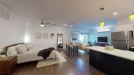Located on John Deshields Boulevard in Bentonville, Arkansas, this single-floor studio is thoughtfully designed to maximize an open, flexible layout. The living and sleeping areas flow seamlessly together, connected by rich hardwood-style flooring that runs throughout the space. A ceiling fan overhead and carefully placed track and pendant lighting provide both functional illumination and a warm ambiance.

The kitchen is a standout feature, showcasing a bold two-tone design with dark lower cabinetry contrasted by crisp white upper cabinets and white quartz countertops. A generous central island offers additional prep and dining space, while vibrant yellow pendant lights add a striking accent. Stainless steel appliances, including a refrigerator, range, and dishwasher, complete the well-equipped kitchen.

The living area benefits from a large sliding glass door that floods the space with natural light and provides access to an outdoor patio with views of the surrounding neighborhood. The bathroom features a floating dark vanity with a backlit mirror, white quartz surfaces, and clean tile flooring. An in-unit stacked washer and dryer is conveniently positioned nearby, adding practical everyday functionality to this polished, contemporary space.