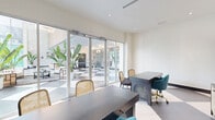 Located at 1625 Northwest 20th Street in Miami, FL, this thoughtfully designed single-floor space offers a compact yet highly functional layout ideal for a professional office or leasing suite. The interior is defined by a striking black-and-white checkerboard tile floor that extends seamlessly from the workspace into the adjacent lobby area, creating a bold visual continuity throughout. Floor-to-ceiling glass walls and automatic sliding glass doors flood the space with abundant natural light while providing clear sightlines into the grand, light-filled lobby beyond, which features tall tropical bird-of-paradise plants and an elegant chandelier. The workspace is furnished with dark wood desks paired with a mix of woven rattan chairs and plush teal velvet seating accented with gold-tone hardware, striking a balance between sophistication and tropical warmth. Two matching rattan-door storage cabinets anchor the rear wall beneath a pair of vibrant framed artwork pieces depicting lush monstera leaves on a pink background, flanked by brass wall sconces that cast a warm, inviting glow. Recessed ceiling lighting throughout ensures consistent illumination. A private alcove area with its own desk and natural light from a full-height side window provides a more intimate workspace within the suite. The overall ambiance is modern, vibrant, and Miami-inspired.