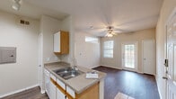 This single-story residence is thoughtfully arranged to maximize functionality across its living areas. The kitchen serves as a central hub, designed in a galley-style configuration with white cabinetry accented by warm wood-tone trim, laminate countertops, a double-basin stainless steel sink, and white appliances including a refrigerator and electric range. Track lighting overhead provides direct task illumination, while an electrical panel is conveniently accessible within the kitchen corridor. A peninsula counter connects the kitchen to the adjoining living and dining area, which features durable wood-look flooring, a ceiling fan with light kit, and a glass-paned exterior door that invites natural light from outside. Windows fitted with blinds provide additional brightness throughout the common areas. The bedrooms are carpeted for comfort, each equipped with ceiling fans. One bedroom includes a built-in shelving unit and desk nook, offering practical storage and workspace. A transom-style interior window adds an architectural detail between spaces. The bathroom is cleanly finished with a tub and shower combination, a vanity with storage, and matching wood-tone cabinetry, continuing the cohesive design found throughout the home.