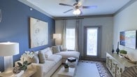 This single-floor space centers around a thoughtfully designed living area that balances comfort and style. The room is anchored by a bold blue accent wall, which contrasts beautifully with the neutral gray tones found throughout the space. A large, abstract canvas artwork adorns the accent wall, adding an artistic focal point to the room.

The living area features a generous white sectional sofa dressed with layered throw pillows, paired with a clean-lined white coffee table resting atop a geometric patterned area rug over hardwood-style flooring. A ceiling fan with an integrated light fixture is centrally mounted, offering both function and a finishing touch to the space.

Natural light flows in through a double window dressed with white blinds and floor-length curtain panels, complemented by a glass-panel entry door that further brightens the room. A floor lamp beside the sofa provides warm ambient lighting for evening use.

Crown molding adds architectural detail along the ceiling line, lending the space a polished, refined character. Overall, the living area presents a cohesive, modern aesthetic with a welcoming and well-lit atmosphere.