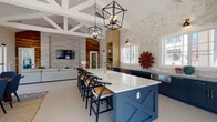 This single-story community clubhouse is thoughtfully designed with a coastal-contemporary aesthetic, combining vaulted, shiplap-clad ceilings with exposed white wooden beams to create an airy, grand atmosphere throughout. Large-format tile flooring flows seamlessly across the main areas, reinforcing the sense of openness.

The kitchen serves as a central focal point, showcasing a bold navy island topped with white quartz countertops, navy cabinetry, stone tile backsplash, stainless steel appliances, and built-in open shelving with wine storage. Geometric black pendant lanterns hang overhead, adding an industrial contrast to the warm, coastal palette.

Adjacent to the kitchen, the main lounge features a sectional seating arrangement anchored by a mounted flat-screen TV, with reclaimed wood accent walls adding warmth. A separate dining area accommodates a large wood table beneath a woven rattan globe pendant, surrounded by abundant natural light from oversized windows overlooking the outdoor pool.

A dedicated workspace includes built-in marble-topped desks, blue shiplap accent walls, and open shelving. A private conference room features a coffered slate-blue accent wall and a live-edge wood table. An additional lounge area offers direct pool views through expansive floor-to-ceiling windows.