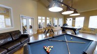 This single-floor recreational amenity space is thoughtfully designed to serve as a vibrant social hub. The room immediately impresses with its soaring vaulted ceilings, which lend an open, airy feel to the entire area. Large windows and glass-paneled doors line the walls, flooding the interior with generous natural light and providing views into adjacent spaces.

The main game room is anchored by a full-size pool table at its center, complete with cues and a racked set of billiard balls ready for play. Overhead, a row of industrial-style pendant lights hangs low, casting focused illumination across the table. Nearby, a foosball table offers additional entertainment, while an air hockey table provides yet another option for friendly competition. A comfortable leather armchair is positioned along one side, offering a place to relax between games.

The flooring throughout is a polished concrete, adding a modern, low-maintenance aesthetic that complements the warm yellow-toned walls. French doors connect this game room to adjacent areas, which appear to include a lounge or business center space, enhancing the overall flow and functionality. The combination of entertainment options, generous proportions, and welcoming design makes this a well-equipped and inviting communal gathering space.