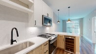 This thoughtfully designed single-floor residence combines contemporary finishes with functional living spaces. The open-concept kitchen serves as the heart of the home, showcasing a two-tone cabinetry scheme that pairs crisp white upper cabinets with warm natural wood lower cabinets. Sleek white quartz countertops run the full length of the kitchen, complemented by a textured scallop-patterned tile backsplash. A full suite of stainless steel appliances includes a gas range, over-the-range microwave, and refrigerator, while a dedicated wine cooler sits at the kitchen island. Stylish pendant lights with mixed metal accents hang above, adding visual warmth. Rich hardwood flooring extends through the kitchen and living area, drawing the eye toward large windows that usher in natural light. The bedroom features plush wall-to-wall carpeting, a ceiling fan, and windows overlooking neighboring structures. The bathroom is elegantly finished with a wide dual-sink vanity in matching natural wood and white quartz, matte black fixtures, a large framed mirror, and a walk-in shower clad in herringbone tile. An adjacent laundry closet houses a stacked GE washer and dryer unit alongside wire shelving for storage, offering a highly practical and complete living environment.