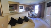 This thoughtfully designed single-floor residence showcases an open-concept layout that effortlessly connects the living, dining, and kitchen areas into one cohesive, welcoming space. The living room anchors the layout with plush, neutral-toned seating accented by dark throw pillows, set atop soft wall-to-wall carpeting that adds warmth underfoot. Adjacent to the living area, the dining space features a stylish round table surrounded by elegantly upholstered chairs, bathed in natural light from a large window framed by rich, floor-length drapery. A decorative sunburst wall accent serves as a striking focal point between the two areas, adding an artistic touch to the warm, earthy color palette throughout. A pendant light fixture overhead lends an intimate ambiance to the dining space. The kitchen is defined by deep, espresso-toned cabinetry, stainless steel appliances, and a granite-topped breakfast bar that serves as both a functional workspace and a natural divider between the kitchen and living areas. Crown molding along the ceiling adds a refined architectural detail, tying the entire space together with a polished, cohesive finish.