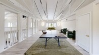 Located at 7912 Olde English Rd in Saint Louis, MO, this two-story property delivers a striking blend of architectural grandeur and recreational versatility across a generously sized footprint. The main level commands immediate attention with its soaring vaulted ceiling supported by exposed white-painted timber trusses, creating a dramatic sense of volume in the living room. A floor-to-ceiling stone fireplace serves as the room's focal point, rising the full height of the wall beneath the cathedral roofline. White French doors, multiple windows, and a chandelier contribute to the bright, airy atmosphere, while dark hardwood floors provide contrast to the all-white architectural elements. A staircase leads to the upper level, which is rimmed by an open balcony railing overlooking the main floor below. The upper level offers light-washed hardwood flooring, exposed beam ceilings, and multiple distinct zones including a spacious game room equipped with a full-size ping pong table, comfortable lounge seating, and an indoor putting green complete with multi-hole turf layout and red flags. Natural light filters through large windows throughout, reinforcing the clean, modern aesthetic that defines this uniquely appointed space.
