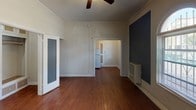 This single-floor space offers a straightforward and functional layout spread across a compact footprint. The main living area is immediately notable for its elegant arched windows that invite natural light, complemented by rich hardwood flooring that flows throughout the unit. Crown molding lines the ceilings, adding a classic architectural touch, while a ceiling fan provides year-round comfort. The walls feature bold, contrasting dark blue accent panels framed in white trim, lending the space a distinctive character. An opening from the living area leads directly into the kitchen, which is efficiently arranged to make the most of its narrow footprint. The kitchen is equipped with a full-size refrigerator and a gas range, providing essential cooking amenities. The bathroom is accessible via a set of double doors and features a full bathtub with wall tile surround, a window for natural ventilation, a pedestal sink, and hardwood flooring that continues from the rest of the unit. Throughout the space, the consistent use of hardwood floors, crown molding, and natural light from multiple windows creates a cohesive and warm ambiance.