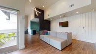 Located on McCormick Street in North Hollywood, this contemporary communal lounge showcases a thoughtfully designed single-floor layout that combines open-plan living with distinct functional zones. The space is immediately defined by its dramatic double-height ceilings, which lend an airy, expansive feel throughout. Warm hardwood flooring runs seamlessly across the entire area, grounding the modern aesthetic.

The central lounge area features a generously sized modular sectional sofa in neutral tones, accented with vibrant pink and teal throw pillows, positioned in front of a textured white shiplap accent wall fitted with a wall-mounted flat-screen television. Overhead, a striking sculptural cross-shaped pendant light fixture serves as a bold focal point above the main seating zone.

A dedicated café-style nook is outfitted with a vivid yellow banquette and paired with round bistro tables and contemporary teal chairs. A large-scale urban cityscape mural anchors this seating area, adding an artistic and cosmopolitan touch. Adjacent to this is access to an outdoor patio furnished with bright chartreuse chairs, inviting natural light deep into the interior through full-height glass doors.

A dark brick accent wall provides a striking contrast to the otherwise crisp white palette, while a teal-accented kitchenette area is visible in the background, completing this well-appointed and vibrant communal gathering space.