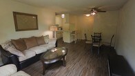 This single-floor space showcases an open-concept design that seamlessly connects the living, dining, and kitchen areas into one cohesive environment. The living area features dark hardwood-style flooring that extends throughout the unit, anchored by a cushioned sofa with accent pillows and an oval glass-top coffee table. A decorative mirror adorns one wall, adding depth to the space. The dining area accommodates a glass-top table with dark wooden chairs, positioned conveniently near the kitchen. A ceiling fan with a light fixture overhead serves both the living and dining zones. The kitchen is defined by white cabinetry, a raised breakfast bar with bar stools, and essential appliances including a microwave and refrigerator, all illuminated by recessed overhead lighting. The layout flows naturally from one area to the next, making the space feel functional and well-connected. Warm, ambient lighting from a table lamp in the living area complements the overall tone of the interior, creating an inviting atmosphere throughout.