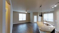 This single-story space in Carrollton showcases an open-concept layout that seamlessly connects the kitchen and living areas. Dark hardwood flooring runs throughout, adding warmth and visual depth to the interior. The living area is well-lit by multiple windows dressed with white blinds, allowing ample natural light to fill the space while maintaining privacy. A ceiling fan with an integrated light fixture anchors the living area, providing both comfort and functionality.

The kitchen area is highlighted by a granite countertop island that serves as a natural dividing point between the cooking and living spaces. Sleek, modern pendant lights hang above, adding a contemporary touch to the design. A dedicated sink is integrated into the island, enhancing the kitchen's utility. Track lighting along the ceiling supplements the pendant fixtures, ensuring the space is well-illuminated.

Neutral-toned walls complement the flooring and finishes throughout, creating a cohesive and inviting atmosphere. A door off the living area provides additional access, contributing to the overall functionality of the layout. The space presents a clean, modern aesthetic suitable for comfortable everyday living.