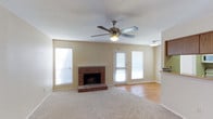 This single-floor home offers a comfortable and well-designed layout that balances functionality with classic charm. The living area serves as the heart of the home, anchored by a traditional brick fireplace with a raised brick hearth — a standout architectural feature that adds warmth and character to the room. Soft carpet flooring covers the main living area, transitioning to smooth hardwood-style flooring toward the adjacent spaces, creating a natural visual distinction between zones. A ceiling fan with a built-in light fixture is centrally mounted, promoting air circulation and providing ambient lighting throughout. Multiple windows dressed with blinds line the walls, inviting generous amounts of natural light while maintaining privacy. A glass-paneled door provides access to the exterior, further enhancing the sense of openness and connectivity to the outdoors. The neutral, light-toned walls offer a versatile backdrop that complements a wide range of interior styles. An adjoining area, visible from the living room, hints at additional functional spaces within the home. Overall, the space presents a cozy yet practical living environment with timeless details throughout.